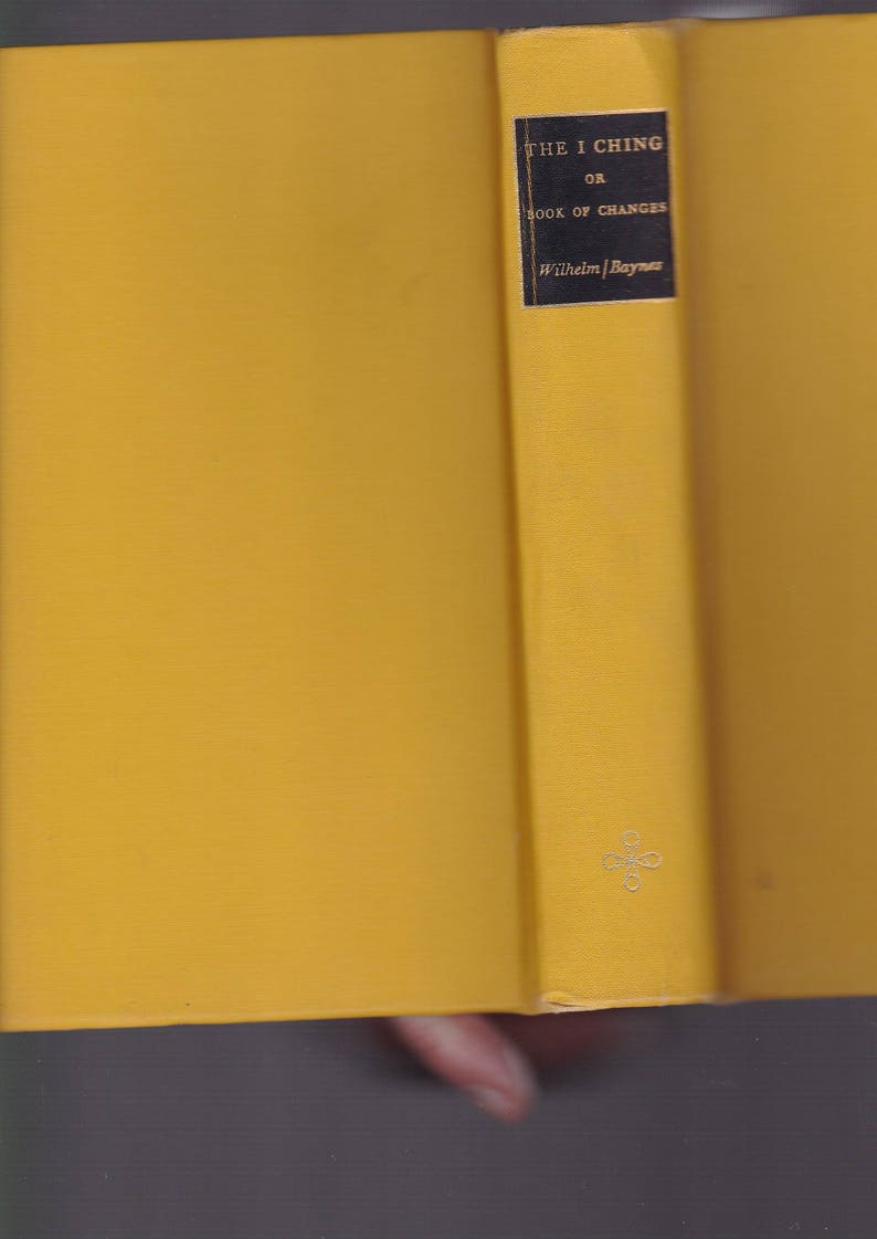 May include: Open book with yellow covers. The spine features a black label with white text: "THE I CHING OR BOOK OF CHANGES" and "Wilhelm/Baynes". A small, silver emblem is visible near the bottom of the spine.