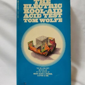 以下が含まれることがあります： トム・ウルフ著『The Electric Kool-Aid Acid Test』のペーパーバック。鮮やかな青い表紙の中央には円形のグラフィックがあり、カラフルな模様の折り紙の容器の中に灰色の立方体が収められている様子が描かれています。