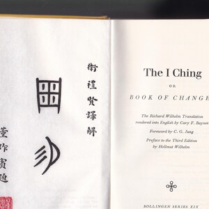 May include: Open book with yellow cover, displaying the title "The I Ching" on the right page. The left page features Chinese characters and a red seal. The book is part of the Bollingen Series XIX.