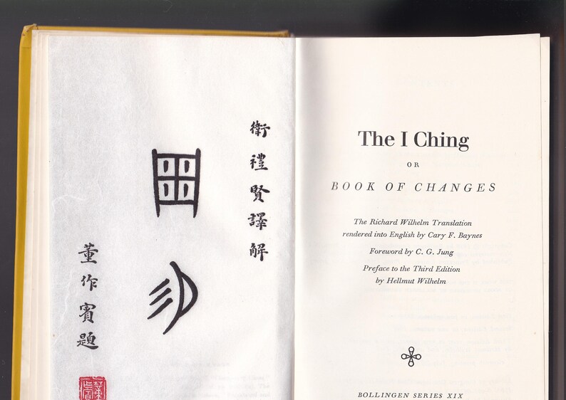 May include: Open book with yellow cover, displaying the title "The I Ching" on the right page. The left page features Chinese characters and a red seal. The book is part of the Bollingen Series XIX.