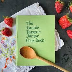May include: A green cookbook titled "The Fannie Farmer Junior Cook Book" with a wooden spoon and fresh strawberries. The book is open on a white cloth with embroidered flowers. The strawberries are red and ripe.