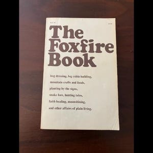 May include: The Foxfire Book, a vintage book with a cream-colored cover and brown text. The title is prominently displayed, with additional text below detailing the book's contents, including hog dressing, log cabin building, and faith healing.