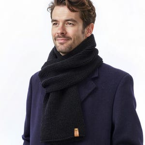 May include: A dark navy blue wool coat worn with a thick black knitted scarf. The scarf features a ribbed texture and a small, rectangular tan leather-colored tag attached to the bottom corner, adding a subtle detail to the winter accessory.