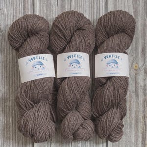 May include: Three skeins of brown yarn with white labels featuring a cartoon sheep wearing a blue hat. The labels read "VOBELLE" and "100% Merino Wool". The yarn is displayed on a light brown wooden surface.