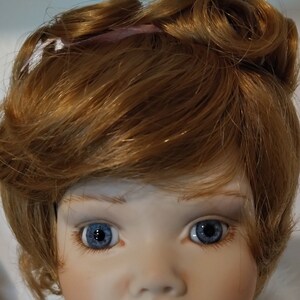 May include: A porcelain doll with a white fur collar and red-brown hair styled in two buns. The doll has blue eyes and a painted smile.