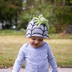 Puede incluir: Un niño pequeño con un sombrero de telaraña gris y negro con una araña verde en la parte superior. El niño lleva una camisa de manga larga azul claro con el texto "No puedo ser bebé hoy" impreso en ella. El niño también lleva pantalones vaqueros azules y zapatos marrones.