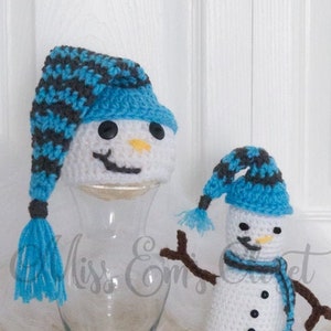 Könnte beinhalten: Zwei gehäkelte Schneemänner, einer groß und einer klein, beide mit blau-grau gestreiften Mützen. Der größere Schneemann sitzt auf einem Glasgefäß, während der kleinere Schneemann auf einer weißen, flauschigen Oberfläche steht.