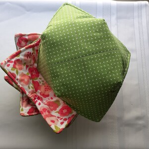 May include: A stack of handmade fabric bowl cozies. The top cozy is green with white polka dots. The interior of the bottom cozy features a floral pattern with pink and red flowers on a white background. The cozies are square and quilted.