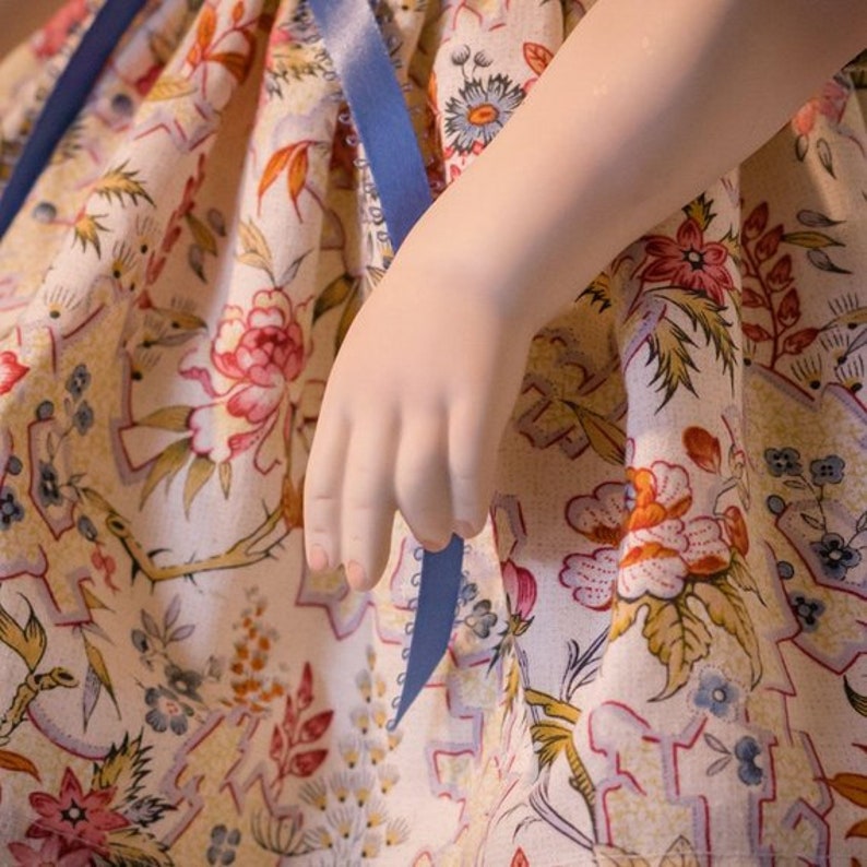 May include: A porcelain doll wearing a white dress with a blue ribbon and a floral pattern. The doll's arm is extended and the hand is visible.