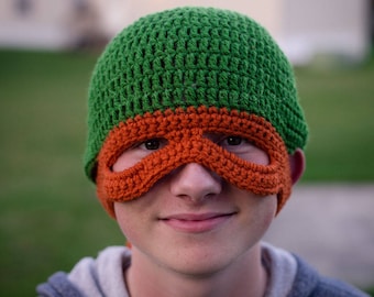 May include: A person wearing a green and orange crocheted hat with a mask. The hat has a green top and an orange mask with eye holes.