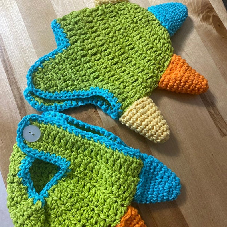 May include: A green, blue, yellow, and orange crocheted dinosaur hat with a button on the top. The hat is made of yarn and has a pointed snout.
