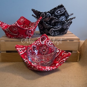 Puede incluir: Cuatro calientaplatos de tela de bandana roja y negra. Los calientaplatos están doblados y expuestos en una caja de madera. Un calientaplatos sostiene un cuenco de vidrio transparente.