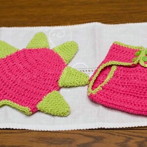 May include: A pink and green crocheted dinosaur hat and diaper set. The hat has a star shape with green spikes and the diaper has a green trim.