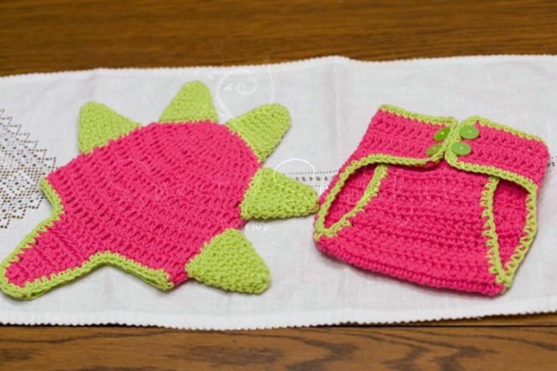 May include: A pink and green crocheted dinosaur hat and diaper set. The hat has a star shape with green spikes and the diaper has a green trim.