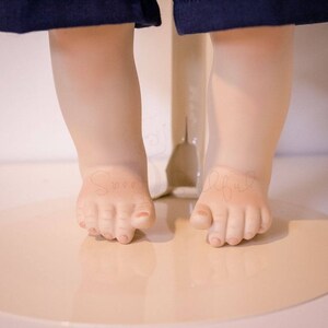 May include: Close-up of a porcelain doll's feet. The doll is wearing blue denim shorts and has bare feet. The feet are posed as if the doll is standing on its toes.