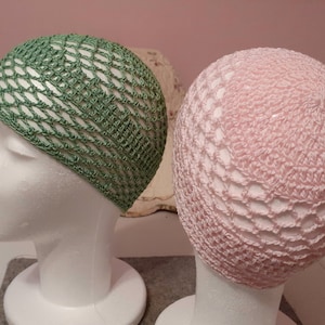 May include: Two crocheted hats, one green and one pink, displayed on styrofoam heads. The hats are made with a lacy stitch pattern.