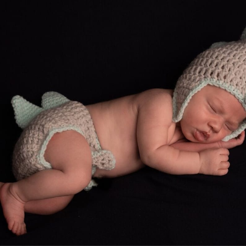 May include: A sleeping baby wearing a light blue and gray crocheted dinosaur hat and matching diaper cover.