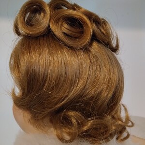 May include: A brown wig with a curled hairstyle. The hair is styled in a vintage updo with two large curls on top and smaller curls around the back of the head.