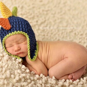 May include: A newborn baby wearing a blue, green, and orange crocheted dinosaur hat. The baby is sleeping on a white fluffy blanket.