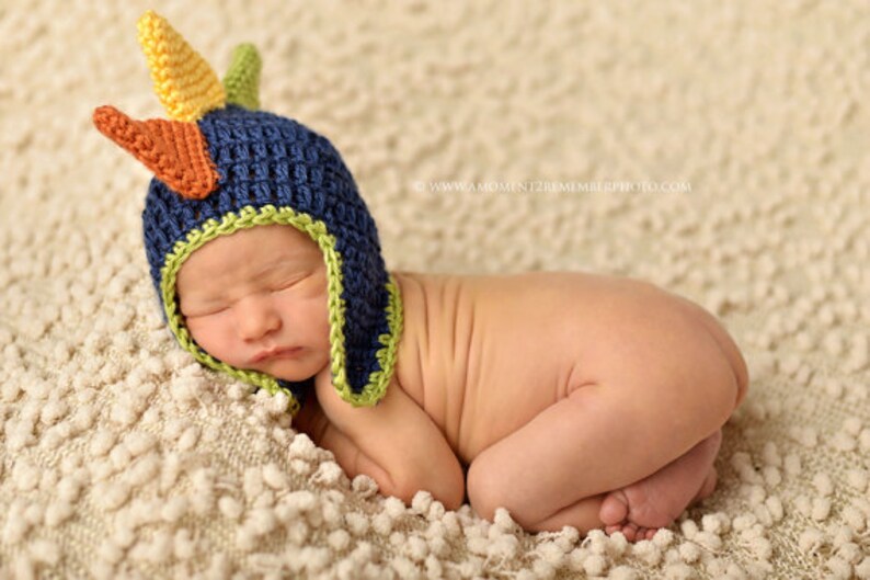 May include: A newborn baby wearing a blue, green, and orange crocheted dinosaur hat. The baby is sleeping on a white fluffy blanket.