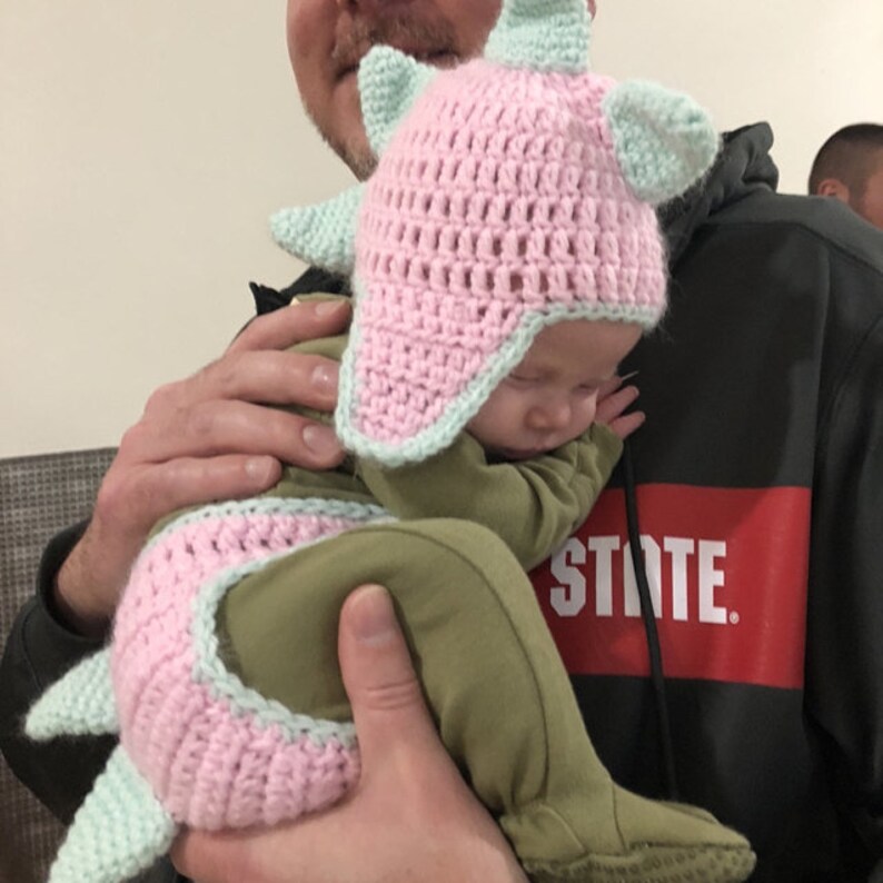 May include: A baby wearing a green and pink crocheted dinosaur costume with a matching hat. The baby is being held by an adult.