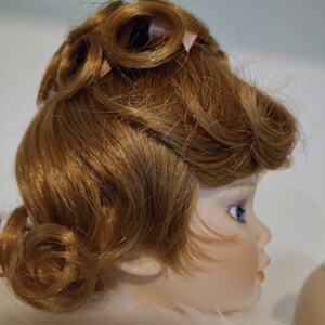 May include: A porcelain doll with a light brown wig styled in a vintage updo with curls. The doll has blue eyes and a painted face.