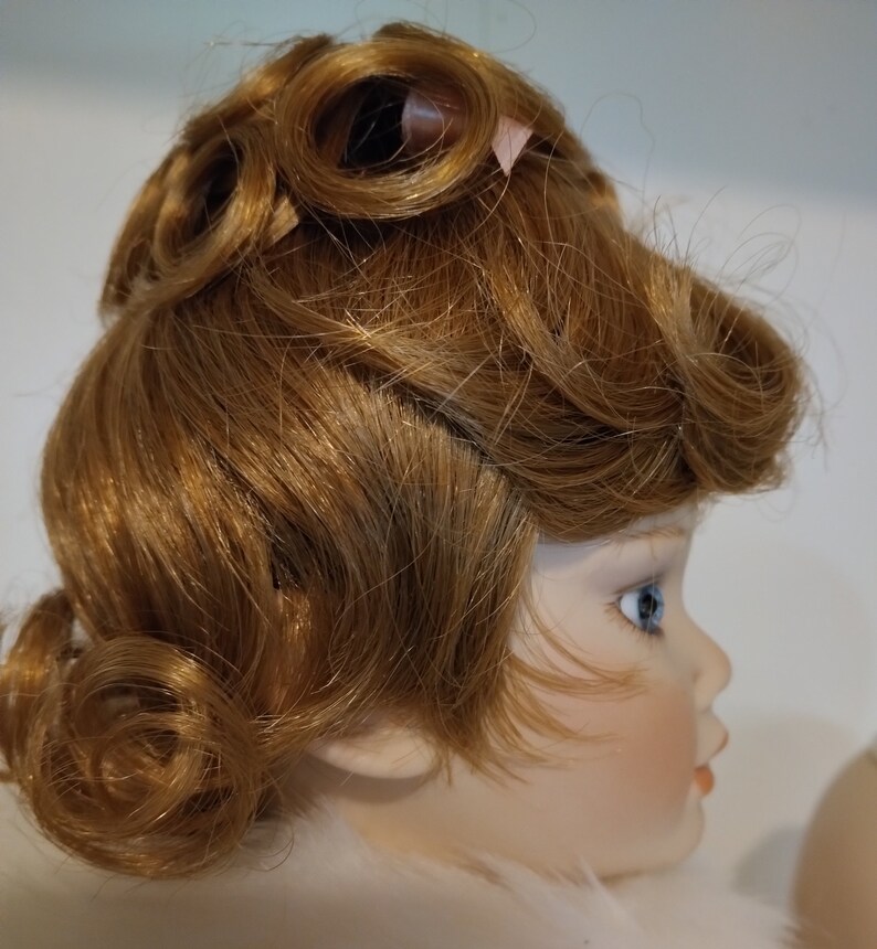 May include: A porcelain doll with a light brown wig styled in a vintage updo with curls. The doll has blue eyes and a painted face.