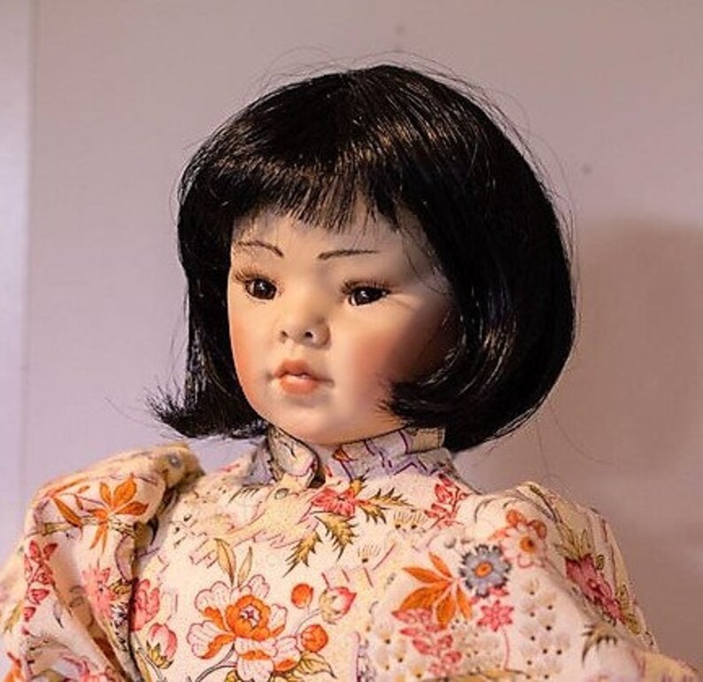 May include: A porcelain doll with black hair styled in a bob, wearing a white dress with a floral pattern.
