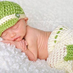 May include: A newborn baby wearing a green and white crocheted hat and matching diaper cover. The baby is sleeping on a white fluffy blanket.