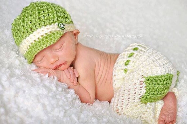 May include: A newborn baby wearing a green and white crocheted hat and matching diaper cover. The baby is sleeping on a white fluffy blanket.