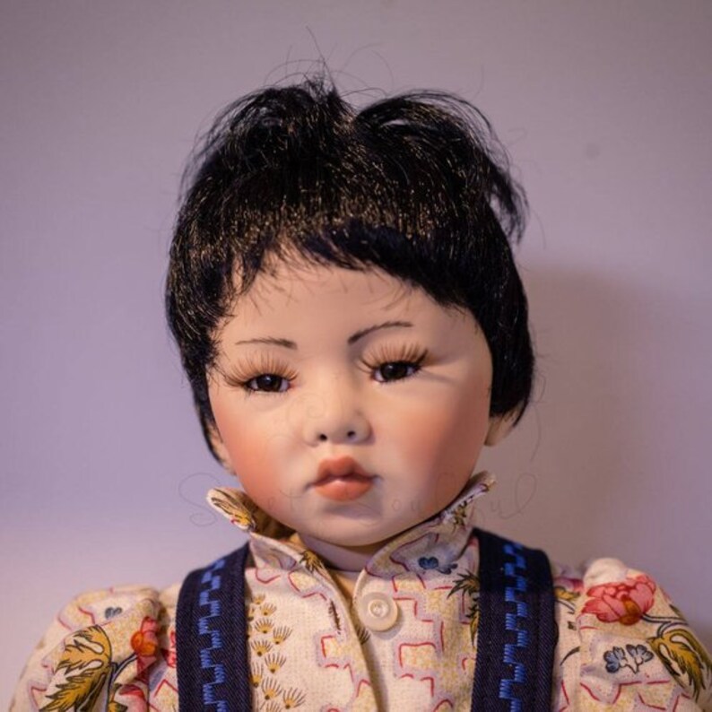 May include: A porcelain doll with black hair and a white and yellow patterned shirt. The doll is wearing blue suspenders and has a serious expression.