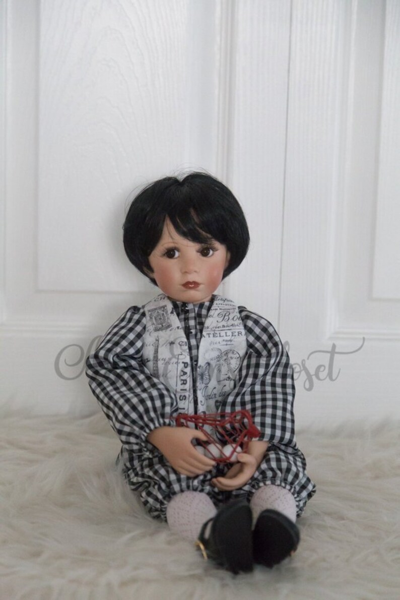 May include: A black and white checkered doll wearing a white scarf with black text and holding a red wire basket. The doll has black hair and brown eyes.