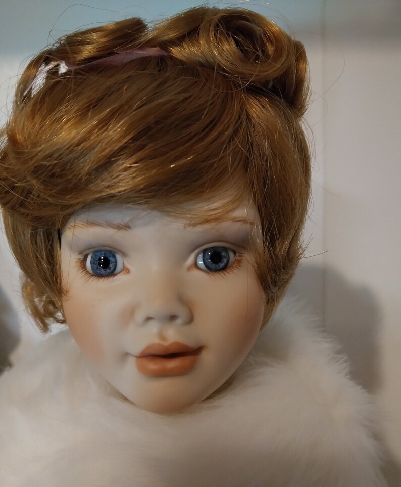 May include: A porcelain doll with a white fur collar and red-brown hair styled in two buns. The doll has blue eyes and a painted smile.