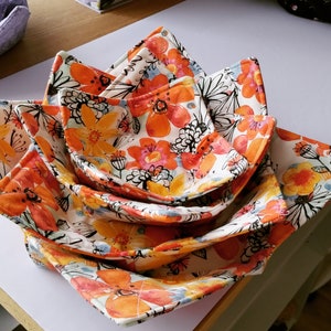 May include: A set of five fabric bowls in a floral pattern with orange, yellow, and white colours. The bowls are nested together and are made of a soft, quilted fabric.
