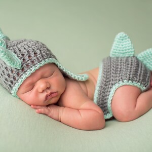 May include: A newborn baby wearing a gray and green crocheted deer hat and matching diaper cover. The baby is sleeping on a green blanket.