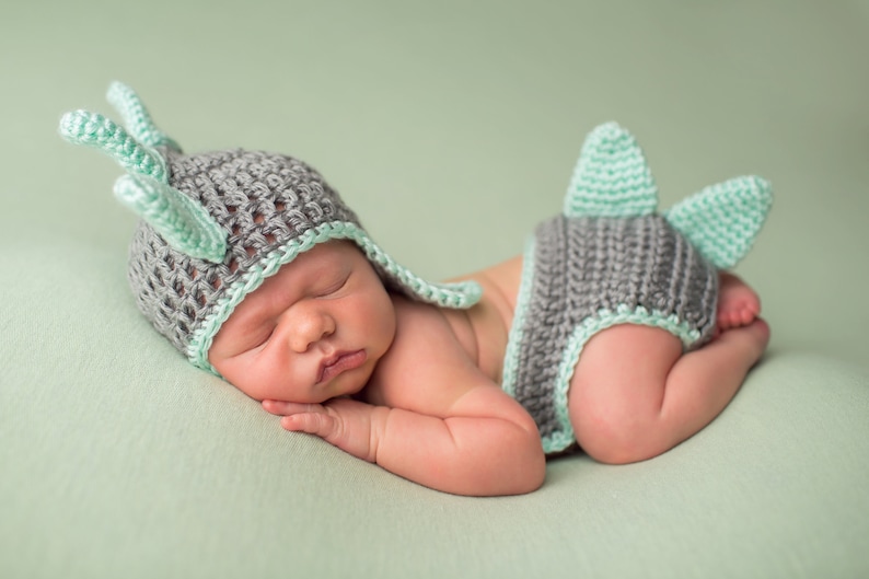 May include: A newborn baby wearing a gray and green crocheted deer hat and matching diaper cover. The baby is sleeping on a green blanket.