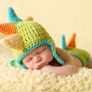 May include: A baby wearing a green, orange, and blue crocheted dinosaur hat and matching diaper cover. The baby is sleeping on a white fluffy blanket.