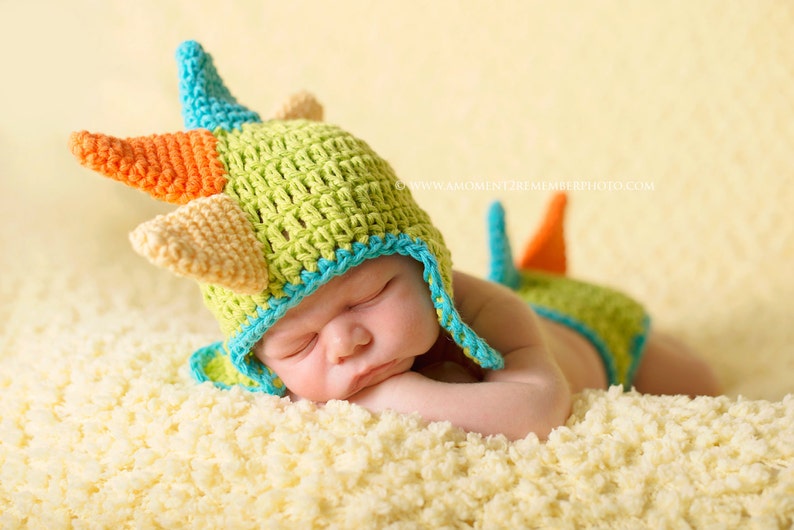 May include: A baby wearing a green, orange, and blue crocheted dinosaur hat and matching diaper cover. The baby is sleeping on a white fluffy blanket.