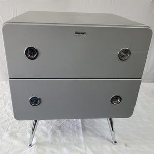 May include: A silver-gray two-drawer nightstand with chrome legs. The drawers have circular, chrome-trimmed handles. The top of the nightstand has a small, rectangular silver plate. The nightstand is on a white surface.