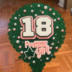 May include: A green glittery cake topper with the number 18 in white with a pink outline. The words "PrettiE Girl" are in pink and white. The topper is decorated with pearls and a green ivy leaf.