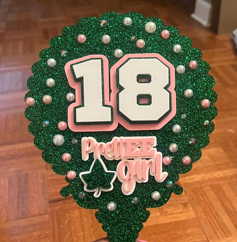 May include: A green glittery cake topper with the number 18 in white with a pink outline. The words "PrettiE Girl" are in pink and white. The topper is decorated with pearls and a green ivy leaf.