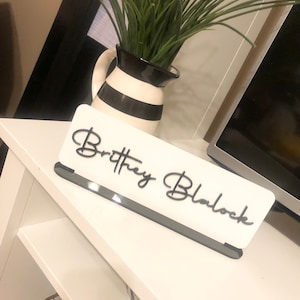 May include: White acrylic desk nameplate with the name "Brittany Blalock" written in black cursive script.