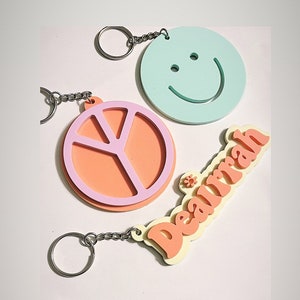 May include: Three colorful keychains with different designs. One keychain is a light blue circle with a smiley face, another is a pink and orange peace sign, and the last is a yellow and orange keychain with the name "Dearrah" written in a retro font.