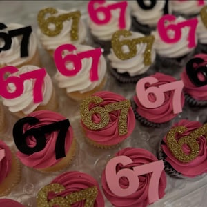 May include: Cupcakes decorated with pink frosting and the number 67 in gold glitter, black, and pink. Some cupcakes have white frosting. The cupcakes are arranged in a clear plastic container.