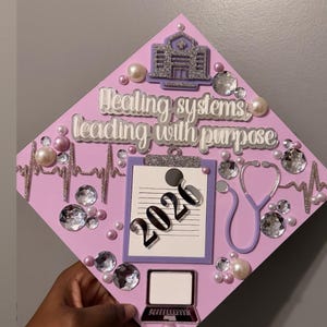 May include: A decorated graduation cap in shades of pink and purple, adorned with rhinestones, pearls, and a stethoscope. The cap features the text "Healing systems, leading with purpose" and the year "2020".