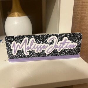 May include: A desk nameplate with the name "Melissa Justice" in a cursive font. The nameplate has a black and white speckled background and a lavender base. A white and gold vase is in the background.