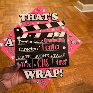 May include: A pink graduation cap decorated with a black and white film clapboard design. The cap reads "THAT'S A WRAP!" and includes the text "Production: Graduation, Director: Laila, 10/26, CHS, 4Yrs". Embellished with rhinestones and pearls.