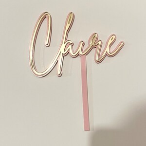 May include: Pink and gold cake topper with the word "Cheers" in a cursive font.