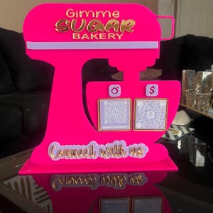 May include: A bright pink acrylic bakery sign shaped like a mixer. The sign features the text "Gimme SUGAR BAKERY" in gold lettering. It includes QR codes for social media and the phrase "Connect with us".