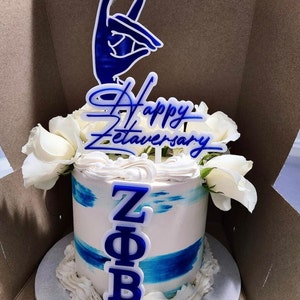 May include: A round cake with white frosting and blue accents, decorated with white roses. The cake is topped with a blue and white sign that says "Happy Zetaversary" and a hand gesture. The letters Z, Φ, and B are also on the cake.